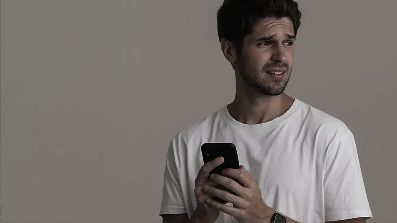 A white man with brown hair holding a mobile phone in front of him with a concerned look on his face against a white wall