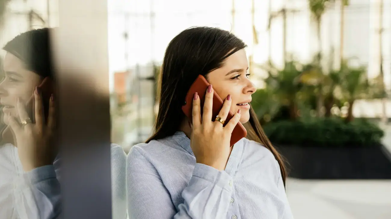 A woman in business casual attire on a mobile phone outside leaning on a mirrored window